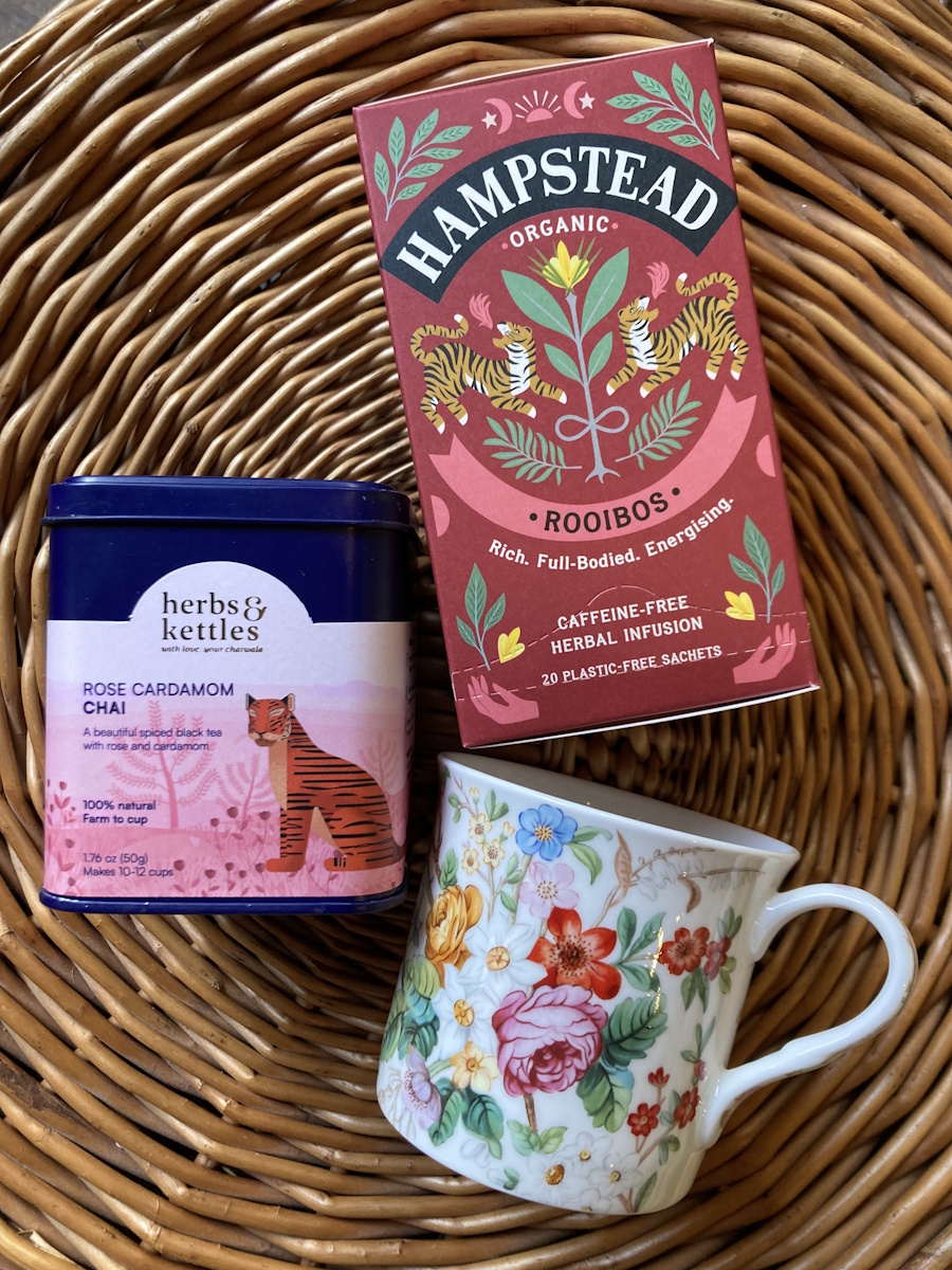 Herbs & Kettles Rose Cardamom Chai tin and Hampstead Organic Rooibos tea box beside a floral teacup, in a wicker basket