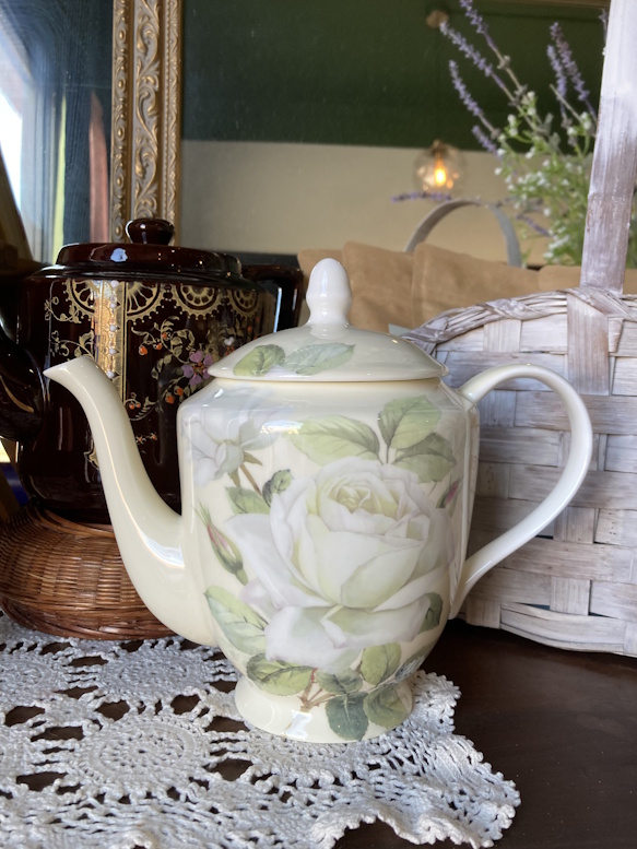 White rose-patterned teapot beside a dark floral teapot on a lace doily