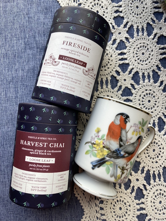 Loose leaf tea tins beside a floral teacup with a bluebird design, on a lace doily